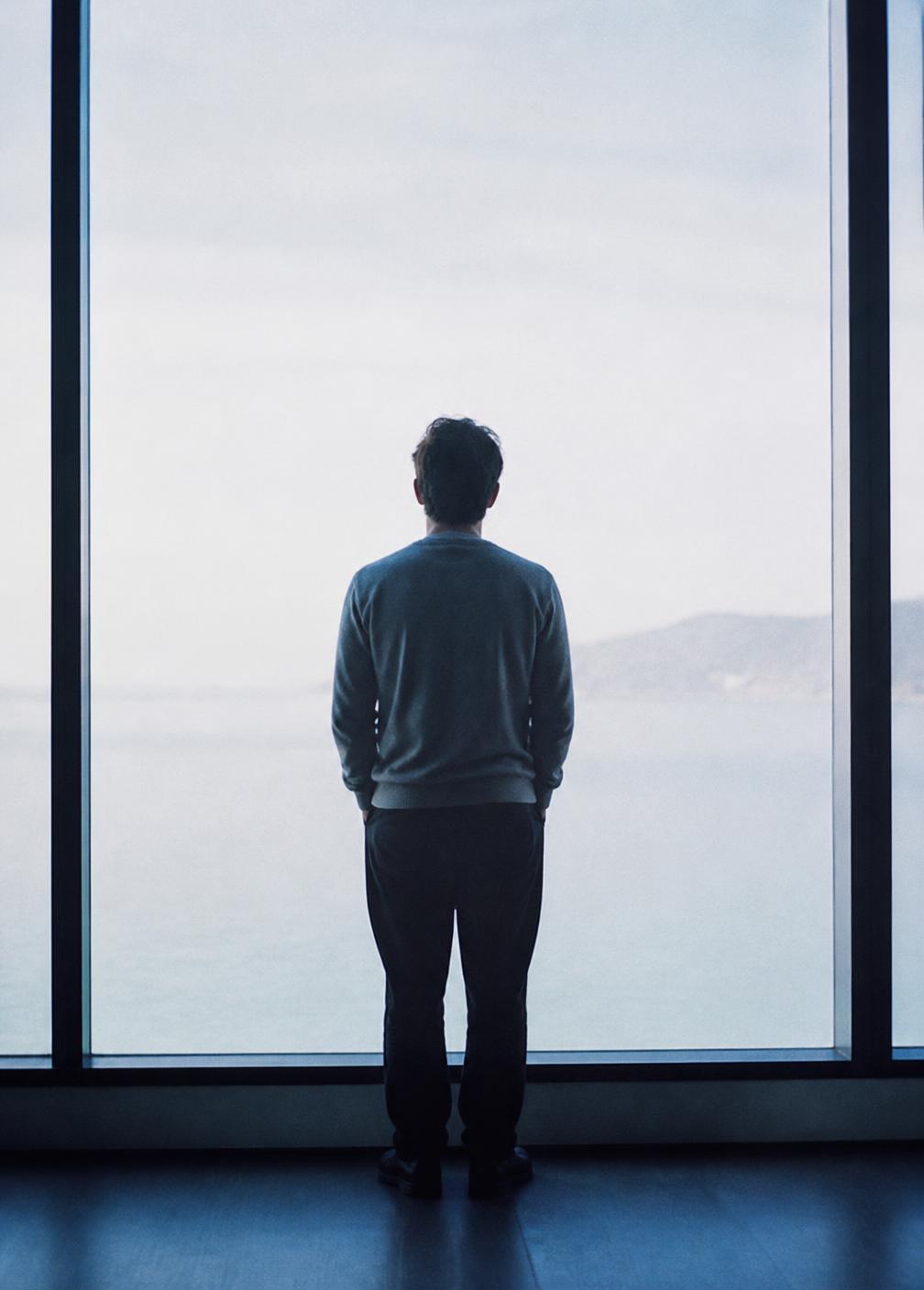 A person standing at a tall window looking out into pale daylight